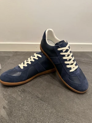 Marinblå sneakers från Maison Margiela - Snygga marinblå sneakers från Maison Margiela med ovandel i mocka och läder, vita skosnören och klassisk gummisula. Skorna har en stilren och tidlös design med låg profil och vita detaljer på insidan. Perfekta för dig som gillar minimalistisk och clean stil. 