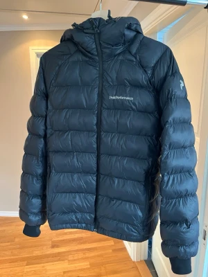 Svart dunjacka Peak Performance - Snygg svart dunjacka från Peak Performance med huva och dragkedja framtill. Jackan har diskret logga på bröstet och vänster ärm, samt ribbade muddar vid ärmslut. Perfekt för kalla vinterdagar tack vare det tjocka dunfodret.