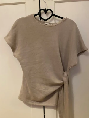 Zara Beige topp med knyt i midjan - Stilren beige topp med korta ärmar och rund halsringning. Toppen har en snygg knyt-detalj i midjan.