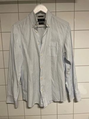 Randig skjorta från Massimo dutti - Säljer en klassisk ljusblå och vit randig skjorta från Massimo dutti. Skjortan har långärmad design, bröstficka och knappar framtill samt på kragen. Perfekt för dig som gillar stilrena och tidlösa plagg.