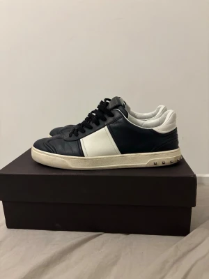 Svarta och vita sneakers från Valentino - Snygga sneakers i svart och vitt läder från Valentino med klassisk låg profil och coola nitar på hälen. Skorna har svarta skosnören, vit panel på sidan och vit sula. Perfekta för dig som gillar stilrena detaljer och exklusiv känsla. Saknar dock en nit på vänster skon men inget man lägger märke till