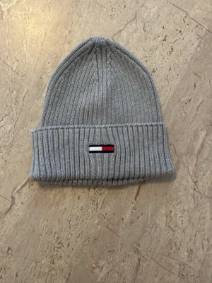 Grå ribbstickad mössa Tommy Hilfiger - Snygg grå ribbstickad mössa från Tommy Hilfiger med uppvikt kant och den klassiska loggan broderad framtill i rött, vitt och blått. Perfekt för kalla dagar och enkel att matcha med olika outfits. Ej använd.