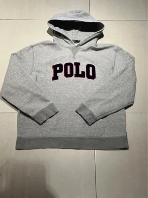 Grå hoodie från Polo Ralph Lauren - Snygg grå hoodie från Polo Ralph Lauren med stor mörkblå POLO-text broderad på bröstet och röda detaljer runt bokstäverna. Klassisk huva och ribbade muddar vid ärmar och nederkant. Tillverkad i mjuk bomullsblandning för skön känsla. Storlek l passar mindre, mått 61 cm längd 58 cm armlängd