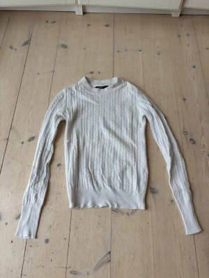 Vit kabelstickad tröja - Kabelstickad vit tröja med rund hals och långa ärmar. Tröjan har ribbade muddar vid ärmslut och nederkant, samt ett klassiskt mönster som ger en stilren look. Perfekt för lager på lager och enkel att matcha med olika outfits. OBS ganska genomskinlig