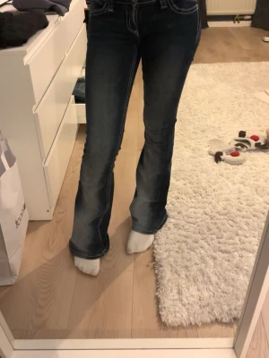 Mörkblå bootcut jeans med brodyr - Säljer ett par mörkblå bootcut jeans med vita kontrastsömmar och snygg broderad ficka bak med paljettdetaljer. Jeansen har låg midja och är tillverkade i stretchigt denim för skön passform. 