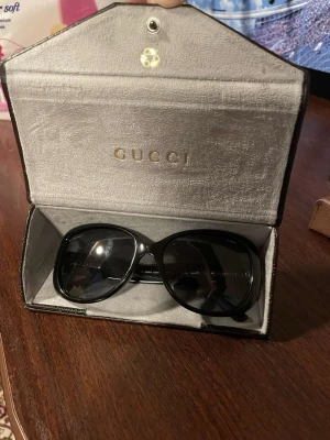 Svarta solglasögon från Gucci - Säljer morsans snygga svarta solglasögon från Gucci med klassisk, fyrkantig form. Kommer i ett exklusivt Gucci-fodral med präglad logga. Perfekta för dig som vill ha en stilren och lyxig accessoar till sommaren.