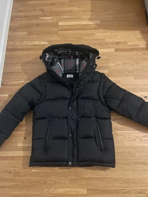 Berberry jacka  - Snygg svart dunjacka från Burberry med avtagbar huva och klassiskt rutigt foder. Jackan har två stora fickor framtill och tryckknappar. Perfekt för kalla vinterdagar och riktigt varm tack vare dunfyllningen.