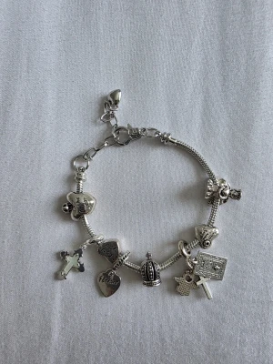 Silverfärgat charmarmband med berlocker - Snyggt silverfärgat armband med flera olika berlocker, bland annat hjärtan med text, ett kors, en krona och en stjärna. Armbandet har en ormkedja och detaljerade hängen som ger en personlig touch. Perfekt accessoar för dig som gillar unika smycken.