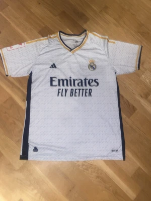 Real Madrid matchtröja Adidas - Snygg vit Real Madrid matchtröja från Adidas med gulddetaljer och mörkblå inslag. Tröjan har klubbmärke på bröstet, Adidas-logga och sponsortryck 'Emirates Fly Better' framtill. Korta ärmar och sportig passform, perfekt för fotboll eller träning.