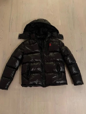 Svart pufferjacka från Polo Ralph Lauren - Säljer en svart pufferjacka från Polo Ralph Lauren med blank finish och avtagbar huva. Jackan har två stora fickor framtill och en röd logga broderad på bröstet. Perfekt för kalla vinterdagar och riktigt snygg streetstil.