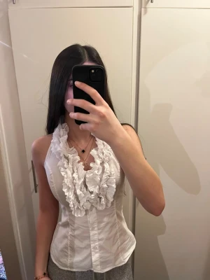 Vit omlott blus med volangdetaljer - Säljer en vit blus med volangdetaljer och omlott-design. Toppen är ärmlös och har en halterneck med knyt i nacken. Den figurnära passformen framhäver siluetten och volangerna ger en trendig look. Perfekt för dig som gillar att sticka ut.