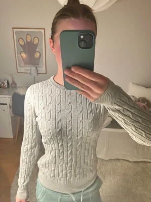 Ljusgrå kabelstickad tröja från Gant - Säljer en ljusgrå kabelstickad tröja från Gant med rund halsringning och ribbade muddar. Tröjan har ett diskret Gant-logo broderat på bröstet och är tillverkad i mjukt material som passar perfekt till jeans eller kjol.