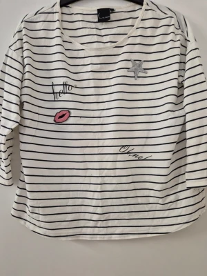 Tröja - Vit tröja med svarta horisontella ränder och coola broderier framtill, som en stjärna, rosa läppar och texten 'hello' och 'oh no!'. Tröjan har rund halsringning och trekvartsärmar. Perfekt för en avslappnad och trendig look. Har några hål och lite gult underarmarna.