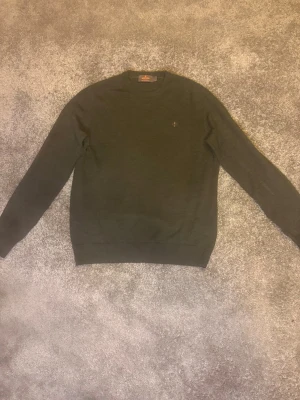 Morris tröja mörkgrön - Mörkgrön stickad tröja från Morris  med rund halsringning och diskret logga broderad på bröstet. Tröjan har ribbade muddar vid ärmslut och nederkant, vilket ger en snygg och klassisk look. Perfekt för dig som gillar stilrena plagg.