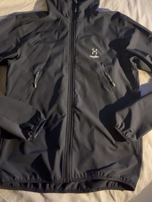 Vindjacka från Haglöfs  - Säljer en svart vindjacka från Haglöfs med huva och två dragkedjeförsedda fickor framtill. Jackan har en stilren design med logotyp på bröstet och är tillverkad i ett lätt och slitstarkt material som passar perfekt för utomhusaktiviteter.