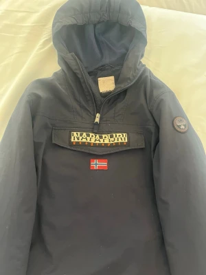 Mörkblå vindjacka från Napapijri - Snygg mörkblå vindjacka från Napapijri med huva och halv dragkedja. Jackan har en stor logotyp på bröstet, norsk flagga och patch på ärmen. Praktisk magficka med dragkedja och meshfoder på insidan. Perfekt för blåsiga dagar. Pris kan diskuteras vid snabb affär.