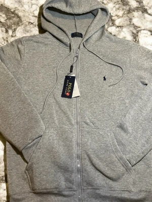 Grå zip hoodie från Polo Ralph Lauren - Snygg grå kofta med dragkedja från Polo Ralph Lauren. Klassisk design med huva och dragsnören, två fickor framtill och den ikoniska mörkblå loggan broderad på bröstet. Tillverkad i mjuk bomullsblandning som känns skön mot huden.