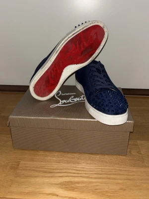 Blå Christian Louboutin sneakers - Snygga mörkblå sneakers från Christian Louboutin med klassisk röd sula och coola nitar på tån. Skorna har en låg profil och är gjorda i mocka med vita gummisulor. Perfekta för dig som vill sticka ut med en exklusiv touch. Väldigt bra skick