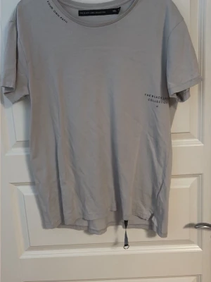 Grå t-shirt The Black Label Collection - Snygg grå t-shirt från The Black Label Collection med diskret tryck på bröstet och text i halsen. Klassisk rund halsringning och korta ärmar. Tillverkad i mjuk bomull för en skön känsla. Perfekt till jeans eller shorts.