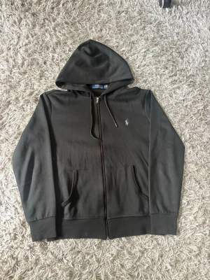Svart hoodie från Polo Ralph Lauren - Klassisk svart hoodie från Polo Ralph Lauren med dragkedja framtill och två fickor. Ikoniska lilla loggan broderad på bröstet. Tillverkad i mjukt bomullsmaterial och har en stor huva med dragsko. Perfekt för en chill och stilren look.