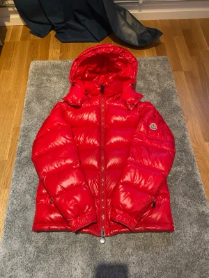 Röd dunjacka från Moncler med luva - Snygg röd dunjacka från Moncler med glansig finish och stor luva. Jackan har dragkedja framtill, fickor med dragkedja och en patch med Moncler-loggan på ärmen. Perfekt för kalla vinterdagar och riktigt trendig look. Självklart äkta,Använd bara fåtal gånger så bra sparsamt använd tveka inte att kontakta om pris eller annat, Mvh Viggo.