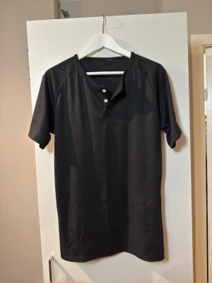 Svart kortärmad topp med knappar - En svart kortärmad topp med v-ringning och tre knappar framtill. T-shirten har en enkel och stilren design med diskret ribbad struktur. Perfekt för dig som gillar en clean look och vill ha något som är lätt att matcha.