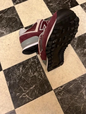 Nya new balance  - Snygga vinröda sneakers från New Balance med klassisk N-logga på sidan. Ovandelen är i mocka och textil, med grå och vita detaljer samt svart yttersula. Skorna har snörning och en bekväm, vadderad häl. Perfekta för dig som gillar streetwear och sportig stil.