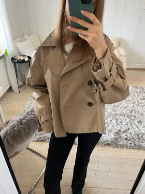 Kort beige trenchcoat med knappar cropped H&M Large - Jättesnygg trenchcoat från H&M i storlek L. Den sitter väldigt fint även på mig som vanligtvis har S. 🥰 Helt slutsåld på hemsidan.   Perfekt jacka nu till våren! Den är i en ljusbeige färg och har klassiska knappar framtill, långa ärmar och stilrena, tidlösa detaljer.   Hör gärna av dig vid frågor eller om du vill ha fler bilder! 😍