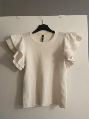 Vit topp med från Golden Days - Supersöt vit topp från Golden Days med volangdetaljer på ärmarna. Rund halsringning och mjukt stickat material som känns skönt mot huden. Perfekt för dig som vill ha en feminin och trendig look.