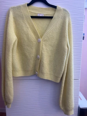 Gul ribbstickad kofta med knappar - Mysig gul ribbstickad kofta  med v-ringning och stora vita knappar framtill. Koftan har långa ärmar och är tillverkad i ett mjukt material som ger en skön känsla. Perfekt att slänga på sig när det är lite kyligare.