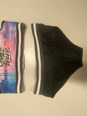 Färgglad bikini med text och ränder - Tvådelad bikini med sportig look. Överdelen har färgglada nyanser i lila, rosa och blått med texten 'Chase the Wave' framtill, samt svarta och vita ränder. Underdelen är svart med matchande ränder i midjan. Snygg och ungdomlig design.