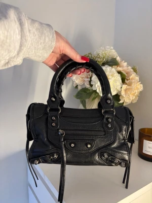 City bag liknande väska - Svart city bag liknande väska i mycket bra skick. Fina detaljer och stängning med dragkedja. Fack framtill med dragkedja. Två mindre öppna fack inuti och ett med dragkedja. Långt band medföljer, längden på bandet går att justera mellan 70 och 120 cm. Mått: 24x14x7,5 cm.