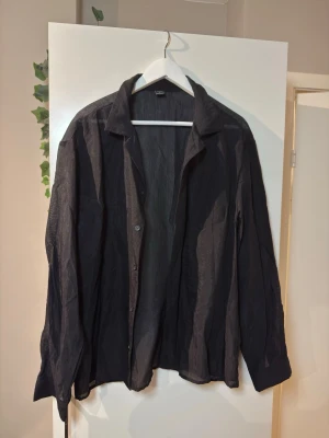 Svart, en liten genomskinlig skjorta - Säljer en svart skjorta med klassisk krage och långa ärmar. Skjortan har knappar framtill och en avslappnad passform som ger en cool och enkel vibe. Perfekt att styla lager på lager eller bära öppen över ett linne.