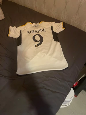 Real Madrid Mbappé matchtröja vit - Snygg vit Real Madrid matchtröja med svarta och gula detaljer. Mbappé och nummer 9 på ryggen, samt klubbmärke och Emirates Fly Better-tryck på bröstet. Tillverkad i lätt och ventilerande material, perfekt för fotbollsträning eller att bära till vardags.