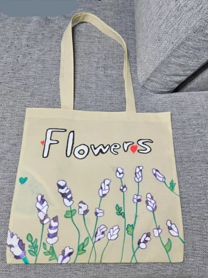 Beige tygkasse med blommotiv - Snygg beige shoppingväska i tyg med handmålade lila och vita blommor samt texten 'Flowers' på framsidan. Väskan har långa handtag och små gröna hjärtan som detaljer. Perfekt för att bära böcker eller matvaror.