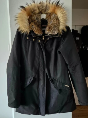 Svart parkas från Woolrich med pälskrage - Snygg svart parkas från Woolrich med stor, fluffig pälskant på huvan i beige och bruna toner. Jackan har dragkedja och knappar framtill, två snedställda fickor och quiltat foder för extra värme. Perfekt för kalla dagar och riktigt stilren look.