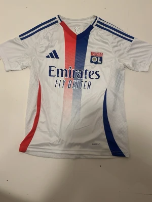 Olympique Lyonnais matchtröja Adidas - Säljer en vit Olympique Lyonnais matchtröja från Adidas med blå och röda detaljer. Tröjan har klubbens logga på bröstet, sponsortryck och klassiska Adidas-ränder på axlarna. Materialet är lätt och andas, perfekt för fotboll eller träning.