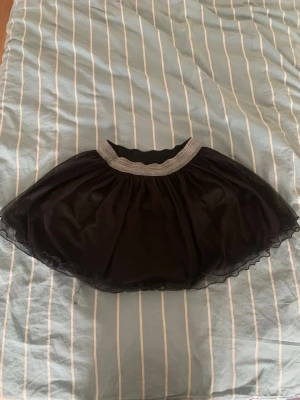 Svart tyllkjol med glittrig resår - Kort svart kjol i tyll med volang och bred glittrig silverfärgad resår i midjan. Den här kjolen har en liten storlek men den passar dig om du är i storlek S och M