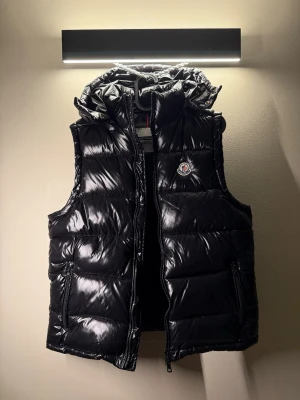 Svart dunväst från Moncler - Snygg svart dunväst från Moncler med glansig finish och klassisk logga på bröstet. Västen har dragkedja framtill, två sidofickor med dragkedja och en avtagbar huva med snörning. Perfekt för lager på lager och streetwear-vibe. Taggar medföljer.