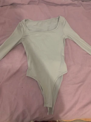 Ljusgrå långärmad body - Säljer en stilren ljusgrå body med långa ärmar och bred, fyrkantig ringning. Bodyn har en enkel och tight passform, perfekt att styla till olika outfits. Materialet känns mjukt och stretchigt, vilket gör den bekväm att bära hela dagen.