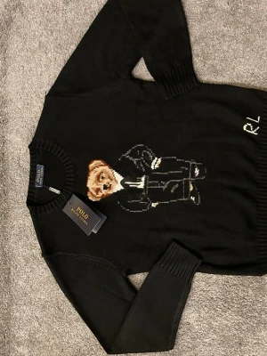 Svart stickad tröja Polo Bear - Svart stickad tröja från Polo Ralph Lauren med ikoniska Polo Bear-motivet framtill. Tröjan har rund hals, ribbade muddar och RL-broderi vid nederkanten. Perfekt för dig som gillar klassisk men lekfull stil. Storlek S.