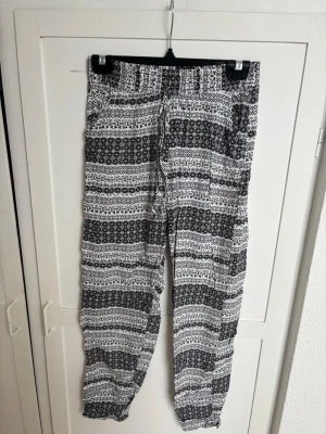 Svarta och vita mönstrade linnebyxor - Säljer ett par luftiga linnebyxor med svartvitt etnoinspirerat mönster. Byxorna har resår i midjan och avslappnad passform, perfekta för chill dagar. Materialet känns lätt och svalt, och de har fickor på sidorna. Perfekt för dig som gillar en avslappnad och trendig stil.