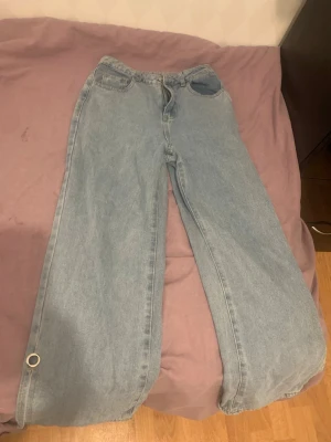 Ljusblåa vida jeans med ringdetalj - Säljer ett par ljusblåa jeans med bred passform och hög midja. Jeansen har klassiska fem fickor och en silverfärgad ringdetalj vid benslutet. Perfekta för dig som gillar en avslappnad och trendig stil.