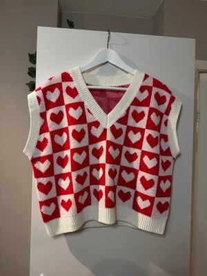 Stickad väst med hjärtan och v-ringning - Säljer en stickad väst i rött och vitt med rutigt hjärtmönster. Västen har breda ribbade kanter, v-ringad hals och croppad passform. Perfekt för dig som vill sticka ut med ett gulligt och trendigt plagg.