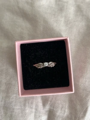 Silverring med vingar och pärldetalj - Unik silverfärgad ring med två detaljerade vingar på sidorna och en liten pärlliknande sten i mitten. Ringen har en elegant och fantasifull design som sticker ut och passar perfekt för dig som gillar smycken med lite extra karaktär.