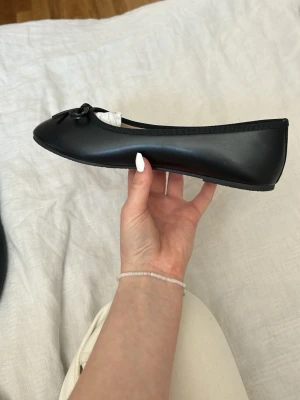 Svarta klassiska ballerinaskor - Stilrena svarta ballerinaskor med rund tå och liten rosett framtill. Skorna har en slät ovansida i syntetmaterial och en tunn, flexibel sula. Dom är köpte utomlands, säljer för dom är för stora nytt skick. Använt en gång
