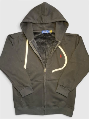 Svart hoodie från Polo Ralph Lauren - Säljer en svart hoodie från Polo Ralph Lauren med dragkedja, vit dragsko och röd broderad logga på bröstet. Hoodien har mjukt foder, ribbade muddar och fickor framtill. Perfekt för en chill och stilren look.