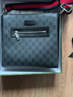 Svart axelväska från Gucci - Snygg svart axelväska från Gucci med klassiskt GG-mönster över hela väskan. Väskan har en justerbar axelrem i rött och svart, en framficka med dragkedja och detaljer i svart läder. Perfekt för dig som vill ha en stilren och trendig accessoar. Jag erbjuder även kvitto och kartongerna från Gucci! Ops, priset går att diskuteras! Butikspris ligger på 14200kr!