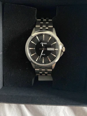 Citizen klocka - Helt ny, oanvänd stilren klocka från Citizen med svart urtavla, silverfärgade index och visare samt datum- och veckodagsvisning. Armband i rostfritt stål med klassisk länkdesign. Perfekt accessoar för dig som gillar en tidlös look.