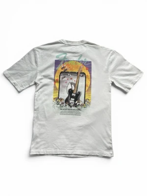 Vit Fender t-shirt med snyggt tryck - Vit t-shirt från Fender med färgglatt tryck på ryggen som visar en elgitarr och förstärkare mot en solnedgång. Framtill finns en liten blå Fender-logga på bröstet. Storleken är XS men är loose fitt och är ganska stor så passa nog bra som S också. Aldrig använd.